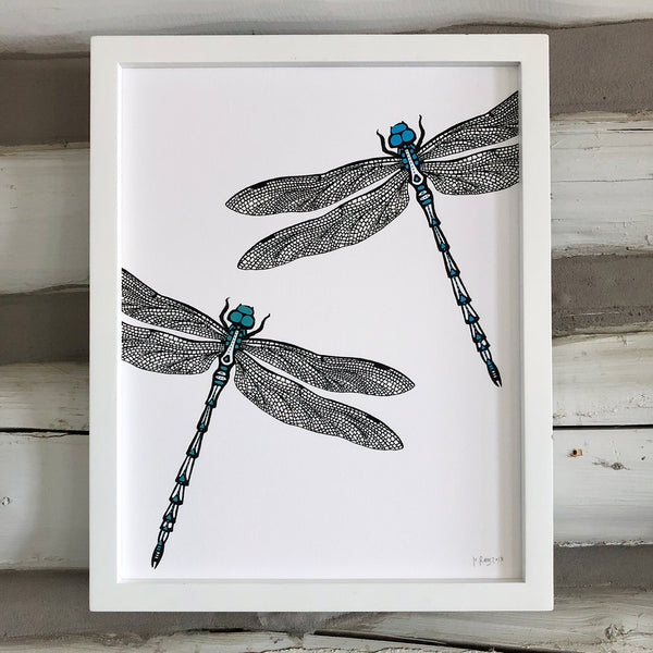 A print of a hand-drawn ink illustration of two dragonflies, one with blue highlights and one with teal accents. Shown in a white frame against a whitewashed log wall. 