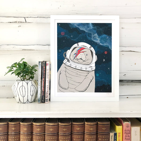 A print of a hand-drawn illustration of a manatee floating in space, wearing an astronaut's helmet with a red and blue lightning bolt over one eye like the iconic image of David Bowie on his Aladdin Sane album cover. Seen in a white frame on a book shelf with a plant and in front of white log walls. 