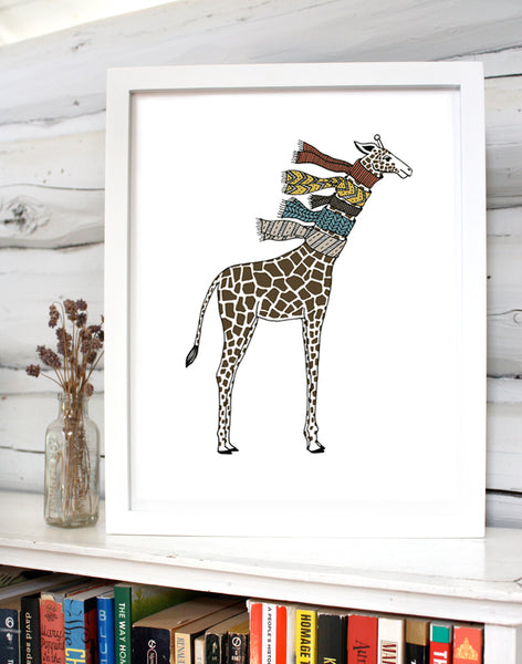 A print of a hand-drawn illustration of giraffe wearing five knit scarves on its neck. Shown in a white frame on top of a bookcase with a small vase of flowers in front of a whitewashed log wall. 