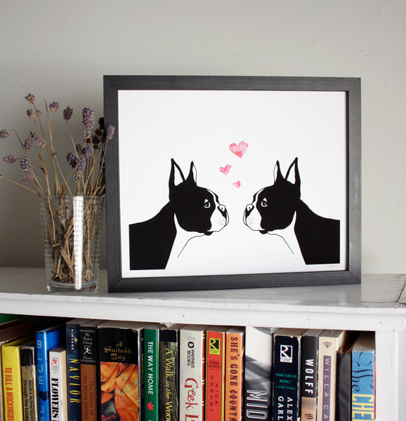 A print of a hand-drawn illustration of two Boston terriers staring into each others eyes with three little hearts rising above their heads. Shown in a black frame on top of a bookcase with a vase of dried lavender next to it. 
