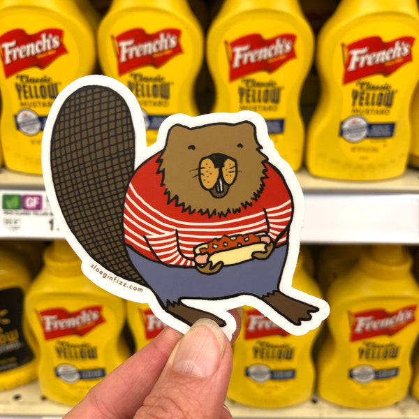 A hand holds up a sticker of an illustrated beaver in a striped red and white sweater holding a hot dog with chili and onions, in front of the French's yellow mustard display at the grocery.