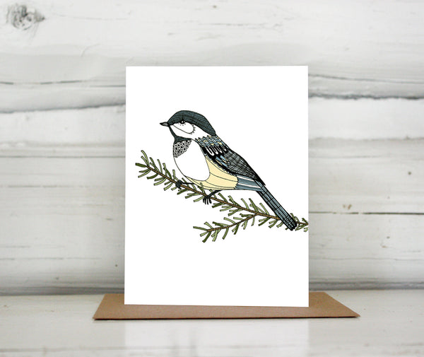 A greeting card with a hand-drawn ink illustration of a chickadee resting on a branch. Shown standing on a Kraft paper envelope in front of a white-washed log wall. 