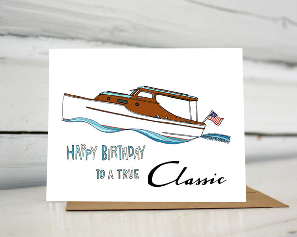 A greeting card with a hand-drawn illustration of a classic Christ Craft boat riding high in the waves with an American flag flying on the stern. A hand-lettered message reads, "Happy Birthday to a true Classic." Shown standing on a Kraft paper envelope in front of a white-washed log wall. 