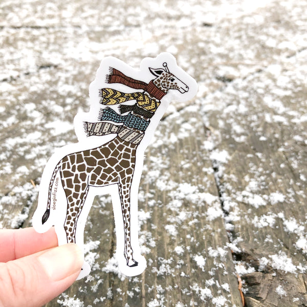A hand holding up an illustrated vinyl sticker of a hand-drawn giraffe wearing five knit scarves on its neck in front of a wood deck covered with little bits of snow.