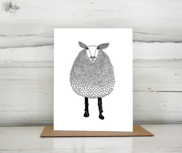 A greeting card with a hand-drawn black and white ink illustration of a sheep. Shown standing on a Kraft paper envelope in front of a white-washed log wall. 