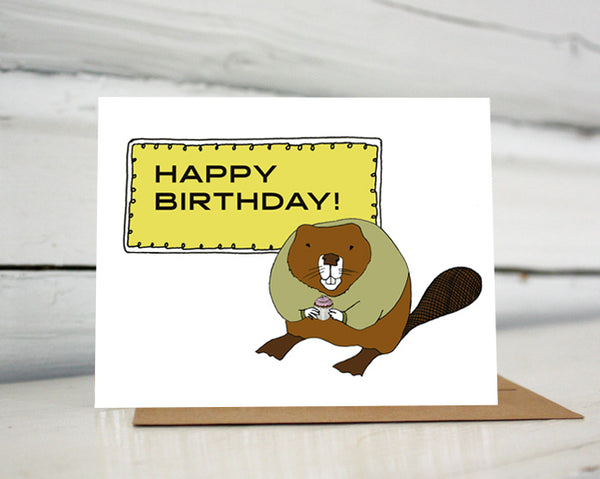 A greeting card with a hand-drawn illustration of a rotund beaver wearing an olive green sweater and holding a cupcake with a pink swirl of frosting standing in front of a sign that reads, " Happy Birthday!" Shown standing on a Kraft paper envelope in front of a white-washed log wall. 
