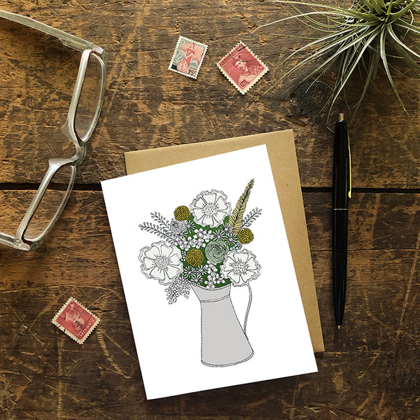 A greeting card showing a hand-drawn illustration of a bouquet of flowers in greys, greens and golds in a silver metal pitcher. Shown with a Kraft paper envelope on a worn wooden surface with reading glasses, stamps, a pen and an air plant. 