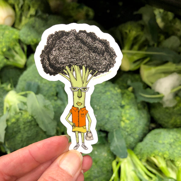 A hand holding a sticker with a hand-drawn ink illustration of a broccoli woman with one hand on her hip, wearing glasses, an orange shirt, yellow skirt and white sneakers, carrying a purse, in front of bunches of real broccoli in the grocery store.