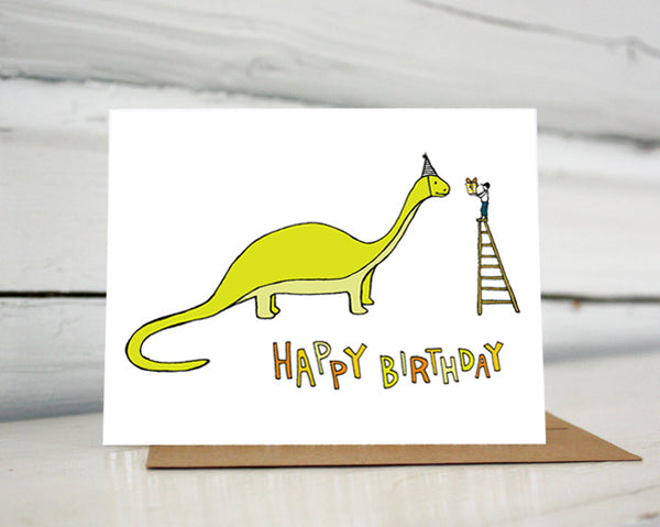 A greeting card showing a hand-drawn illustration of a green brontosaurus wearing a party hat. A small boy stands on top of a ladder handing the dinosaur a wrapped birthday gift. A hand-lettered message reads, "Happy Birthday." Shown standing on a Kraft paper envelope in front of a white-washed log wall. 