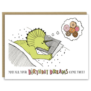A greeting card showing a hand-drawn illustration of a green stegosaurus dinosaur sleeping on moons and stars sheets with his head on two pillows. He is dreaming of a six doughnuts in a variety of colors and flavors. A hand-lettered message reads, "May all your birthday dreams come true." Shown with a Kraft paper envelope on a white background. 
