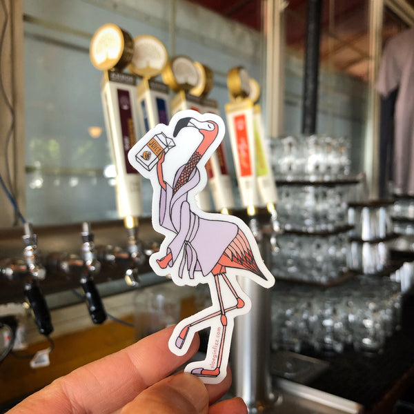 A hand holding up a vinyl sticker of a hand-drawn flamingo wearing a lavender bathrobe and lavender slippers, with its head tipped back to drink from an orange juice carton, in a bar room.