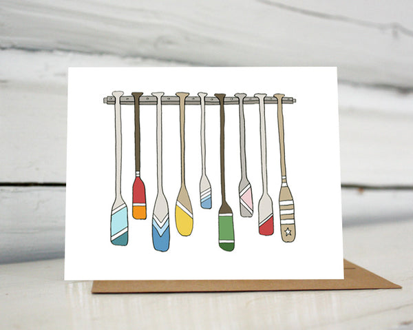 A greeting card with a hand-drawn illustration of a rack full of colorful oars and paddles each decorated with different color and stripes. Shown standing on a Kraft paper envelope in front of a white-washed log wall. 