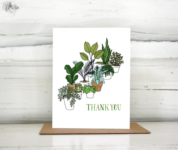 A greeting card showing a hand-drawn illustration of a collection of houseplants. The card has a hand-lettered message reading, "Thank You." Shown standing on a Kraft paper envelope in front of a white-washed log wall. 