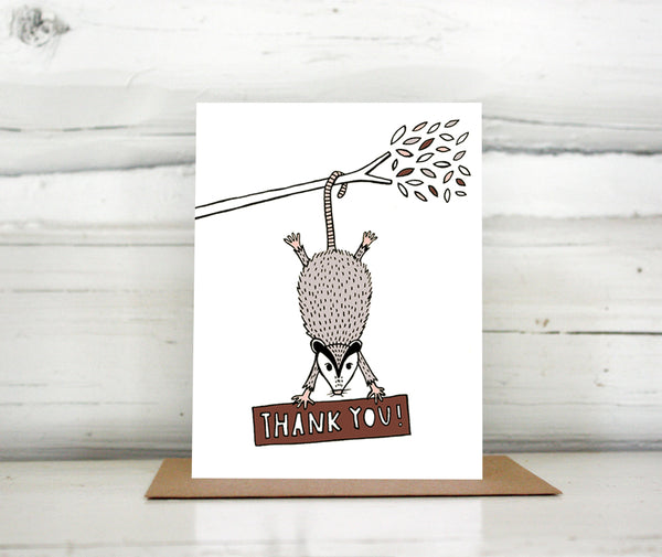 A greeting card showing a hand-drawn illustration of a possum hanging from a tree branch hiding a sign with a hand-lettered message that reads,"Thank you!" Shown standing on a Kraft paper envelope in front of a white-washed log wall. 