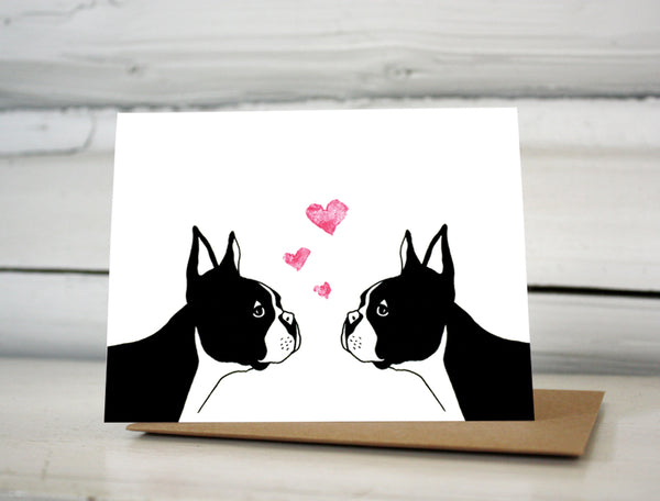 A greeting card with a hand-drawn illustration of two Boston terriers facing each other staring into each others eyes with three little hearts rising above their heads. Shown standing on a Kraft paper envelope in front of a white-washed log wall. 