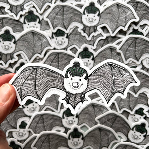Fingertips hold a die-cut sticker featuring a hand-drawn illustration of a cute bat with a little hog nose wearing a knit hat.  