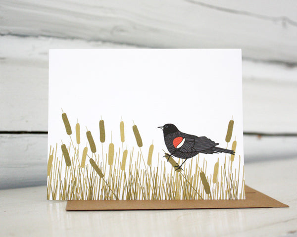 A greeting card showing a hand-drawn illustration of red-winged blackbird resting amongst a swath of cattails. Shown standing on a Kraft paper envelope in front of a white-washed log wall. 