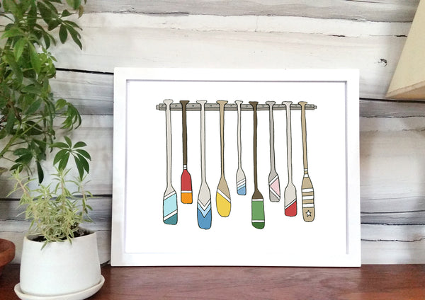 A print of a hand-drawn illustration of a rack full of colorful oars and paddles each decorated with different color and stripes. Shown on in a white frame in front of a whitewashed log wall.