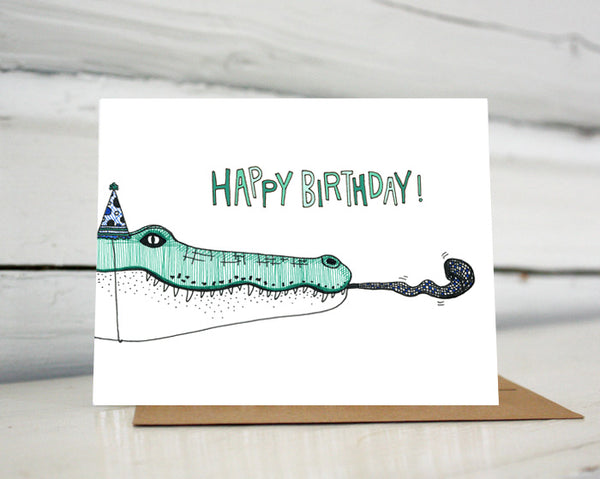 A greeting card showing a hand-drawn ink illustration of a green and white crocodile wearing a polka-dotted party hat and blowing a noisemaker. A hand-lettered message reads, "Happy Birthday!" Shown standing on a Kraft paper envelope in front of a white-washed log wall. 