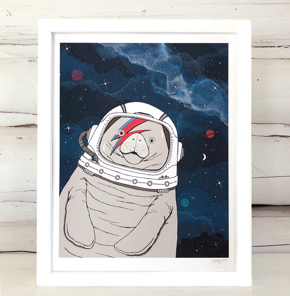 A framed print of a hand-drawn illustration of a manatee floating in space, wearing an astronaut's helmet with a red and blue lightning bolt over one eye like the iconic image of David Bowie, ganging on a white log wall. 