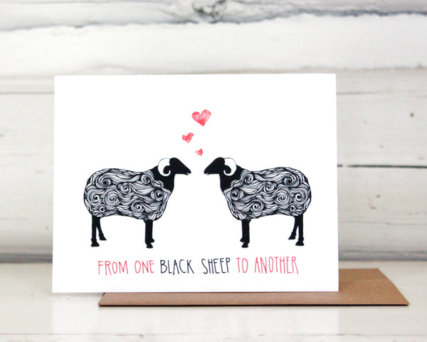 A greeting card with a hand-drawn ink illustration of a pair of black and white sheep with horns facing each other with three hearts over their heads. A hand-lettered message reads, "From one black sheep to another." Shown standing on a Kraft paper envelope in front of a white-washed log wall. 