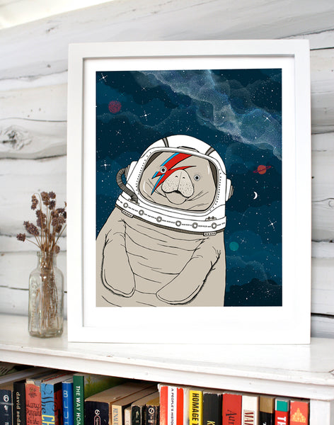 A print of a hand-drawn illustration of a manatee floating in space, wearing an astronaut's helmet with a red and blue lightning bolt over one eye like the iconic image of David Bowie on his Aladdin Sane album cover. Seen in a white frame on a book shelf in front of white log walls. 