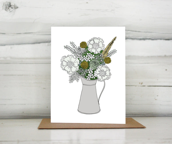A greeting card showing a hand-drawn illustration of a bouquet of flowers in greys, greens and golds in a silver metal pitcher. Shown standing up on a Kraft paper envelope in front of a white-washed log wall. 