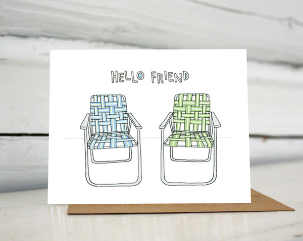 A greeting card showing a hand-drawn illustration of a pair of old-fashioned lawn chairs with webbing and metal frames, one in blues and one in greens. A hand-lettered message reads, "Hello Friend." Shown standing on a Kraft paper envelope in front of a white-washed log wall. 