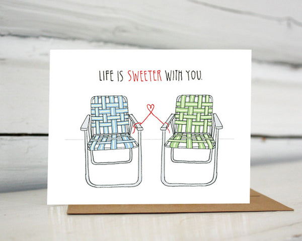 A greeting card showing a hand-drawn illustration of a pair of old-fashioned lawn chairs with webbing and metal frames, one in blues and one in greens. A red thread ties the two chairs together and forms a small heart shape between them. A hand-lettered message reads, "Life is sweeter with you." Shown standing on a Kraft paper envelope in front of a white-washed log wall. 