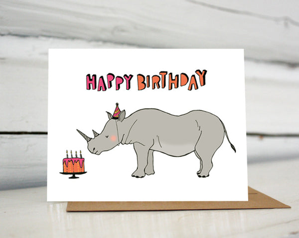 A greeting card showing a hand-drawn illustration of a rhinoceros wearing a pink and black striped party hat that matches stripes on his horns, looking at an orange and pink birthday cake. A hand-lettered message reads, "Happy Birthday." Shown standing on a Kraft paper envelope in front of a white-washed log wall. 