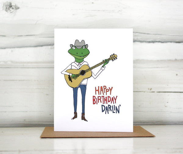 A greeting card showing a hand-drawn illustration of a frog playing a guitar, wearing cowboy boots, a cowboy hat, jeans and a white Western shirt. Shown standing on a Kraft paper envelope in front of a white-washed log wall. 
