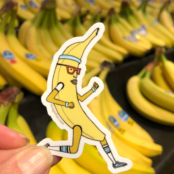A hand holding up a sticker with an illustration of a running banana wearing glasses, sweatbands, knee socks and running shoes  in front of bunches of bananas in a grocery store.