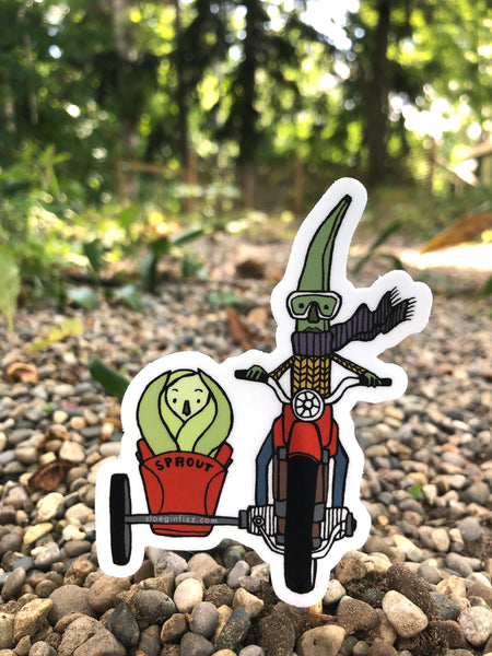 A sticker with an illustration of a green bean wearing goggles, a purple scarf and yellow sweater riding a red motorcycle with a sidecar carrying a Brussel sprout is seen riding along a gravel path in the woods.