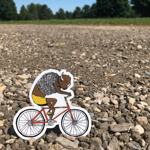 A vinyl sticker with a hand-drawn illustration of a bison riding a red bicycle wearing yellow cycling shorts and yellow sneakers is seen riding along a gravel path with woods in the background. 