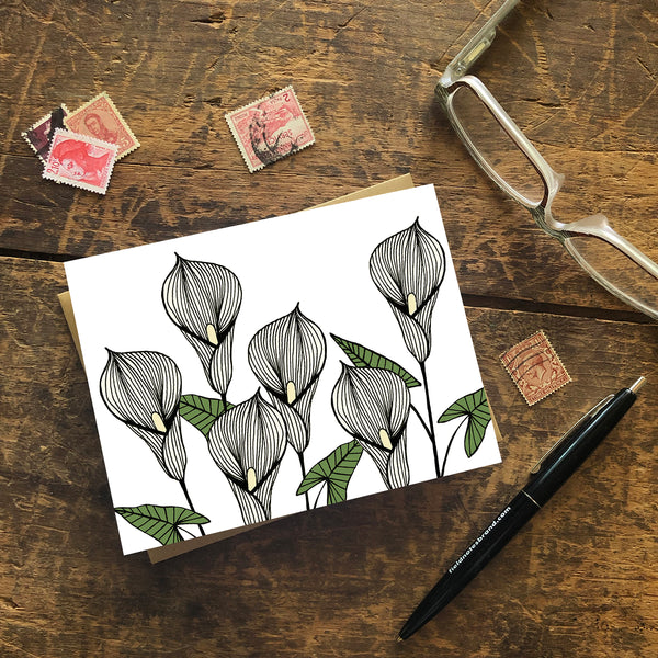 A greeting card with a hand-drawn ink illustration of six pale yellow calla lilies. Shown with a Kraft paper envelope on a worn wooden surface with reading glasses, stamps and a pen. 