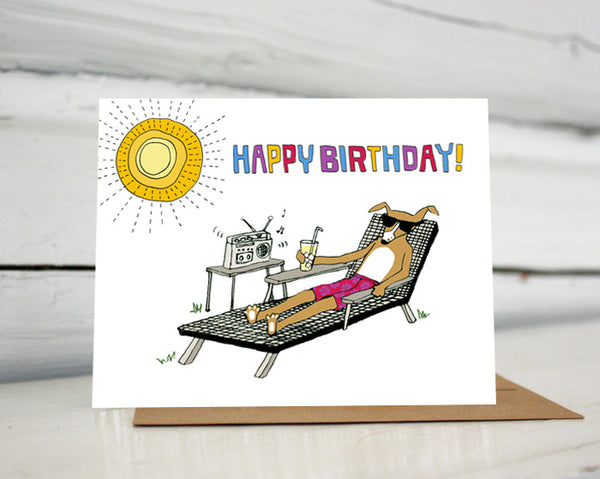 A greeting card with a hand-drawn illustration of a dog in sunglasses and a floral bathing suit sitting in a lounge chair in the yard, drinking a tall glass of lemonade and listening to a boombox with one speaker while the sun shines overhead. A hand-lettered message reads, "Happy Birthday!" Shown standing on a Kraft paper envelope in front of a white-washed log wall. 