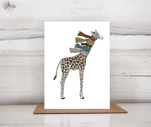 A greeting card showing a hand-drawn illustration of giraffe wearing five knit scarves on its neck. Shown standing on a Kraft paper envelope in front of a white-washed log wall. 