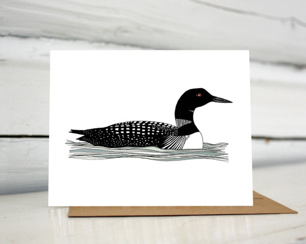 A greeting card showing a hand-drawn illustration of a black and white loon with its signature red eye, floating gently on the waves. Shown standing on a Kraft paper envelope in front of a white-washed log wall. 