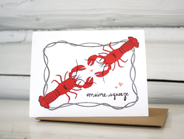 A greeting card with a hand-drawn ink illustration of two bright red lobsters facing each other with hearts around and a frame of nautical roping. The hand-lettered message reads, "Maine Squeeze." Shown standing on a Kraft paper envelope in front of a white-washed log wall. 