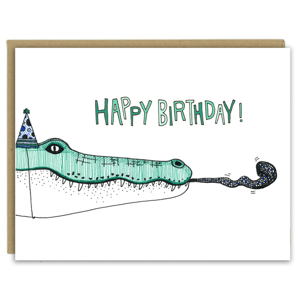 A greeting card with a hand-drawn ink illustration of a green and white crocodile wearing a polka-dotted party hat and blowing a noisemaker. A hand-lettered message reads, "Happy Birthday!" Shown with a Kraft paper envelope on a white background. 
