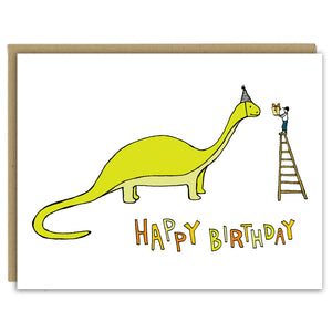 A greeting card showing a hand-drawn illustration of a green brontosaurus wearing a party hat. A small boy stands on top of a ladder handing the dinosaur a wrapped birthday gift. A hand-lettered message reads, "Happy Birthday." Shown with a Kraft paper envelope on a white background. 