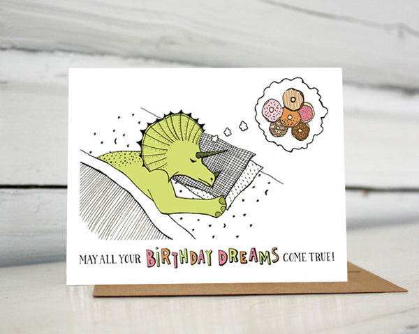 A greeting card showing a hand-drawn illustration of a green stegosaurus dinosaur sleeping on moons and stars sheets with his head on two pillows. He is dreaming of a six doughnuts in a variety of colors and flavors. A hand-lettered message reads, "May all your birthday dreams come true." Shown standing on a Kraft paper envelope in front of a white-washed log wall. 