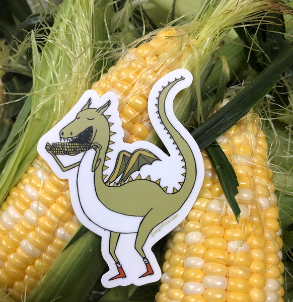 A sticker with a hand-drawn green dragon eating corn on the cob and wearing little red boots seen in front of several ears of corn.