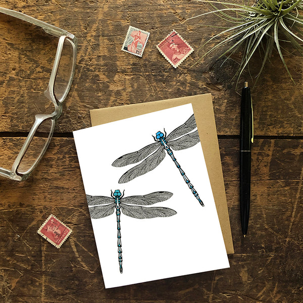 A greeting card showing a hand-drawn ink illustration of two dragonflies, one with blue highlights and one with teal accents. Shown with a Kraft paper envelope on a worn wooden surface with reading glasses, stamps, a pen and an air plant. 
