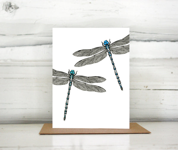 A greeting card showing a hand-drawn ink illustration of two dragonflies, one with blue highlights and one with teal accents. Shown standing on a Kraft paper envelope in front of a white-washed log wall. 