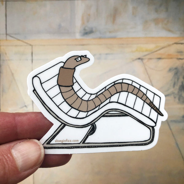 A hand holding up a sticker with a hand-drawn ink illustration of an earthworm reclining on a white mid-century modern lounge chair, in front of a muted abstract background. 
