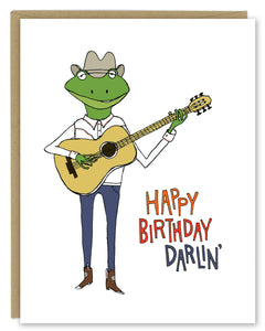 A greeting card showing a hand-drawn illustration of a frog playing a guitar, wearing cowboy boots, a cowboy hat, jeans and a white Western shirt. Shown with a Kraft paper envelope on a white background. 