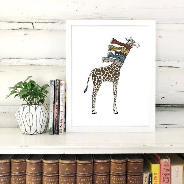 A print of a hand-drawn illustration of giraffe wearing five knit scarves on its neck. Shown in a white frame on top of a bookcase with a potted plant and a few books, in front of a whitewashed log wall. 