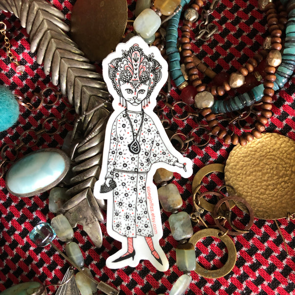 An illustrated die-cut vinyl sticker of a hand-drawn cat decked out in glamorous 1920s style, with a drop waist patterned dress, an elaborate headdress, dangly earrings and a clutch. She has a large pendant around her neck, red stockings and pointy shoes. Seen on a background of various jewelry.