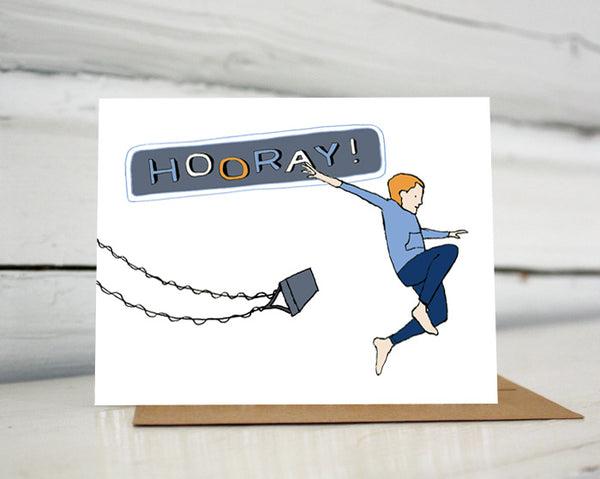 A greeting card showing a hand-drawn illustration of a red-headed boy wearing navy blue pants and a blue gray sweatshirt, arms spread wide, jumping off a high-flying swing. "Hooray!" says the land-lettered message on the card. Shown standing on a Kraft paper envelope in front of a white-washed log wall. 